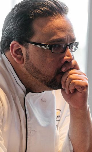 A thoughtful chef wearing glasses, resting his chin on his hand.
