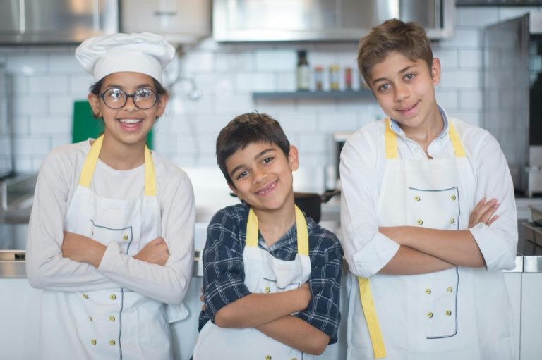 Three children in chef attire pose confidently in a kitchen, smiling with arms crossed.