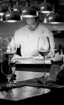A focused chef preparing a dish in a dimly lit restaurant, with wine glasses in the foreground.