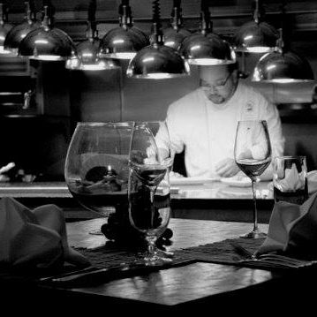 Chef aLEX Rosado preparing a dish in a dimly lit kitchen, with wine glasses in the foreground.