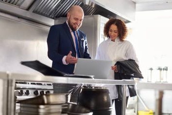 A man in a suit and a woman in a chef's coat discuss something on a laptop in a kitchen.