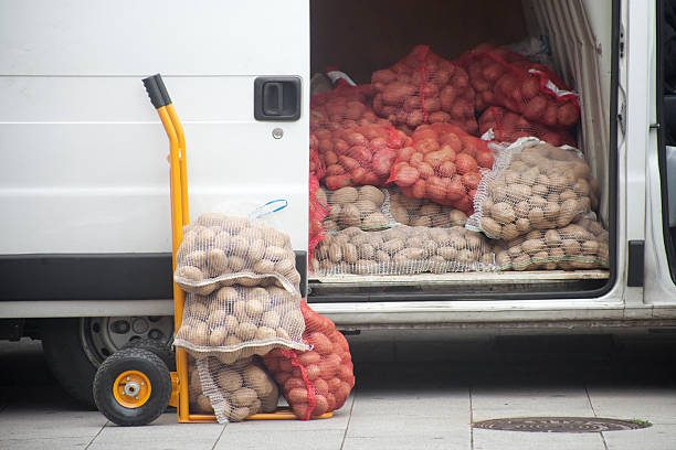A van loaded with bags of red and brown potatoes, with a hand truck beside it.